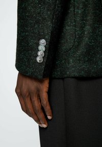 Mörkgrön ullblandad jacka med texturfinish, prydd med fyra metallknappar på ärmen, som bärs tillsammans med en svart tygbyxa.