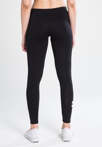 Svart tights med elastisk midjebånd, glatt tekstur, og en hvit logo detalj på underbenet. Brukt med hvite sneakers.