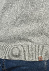Pull en tricot gris avec ourlet côtelé, présentant un design texturé et une petite étiquette en cuir marron en bas.