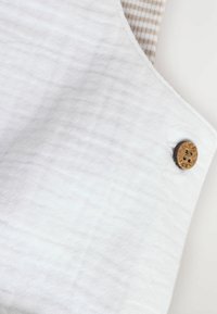Weißer strukturierter Stoff mit einem Holzknopfverschluss. Der Hintergrund besteht aus einem hellbeigen Textil mit feinen Streifen.