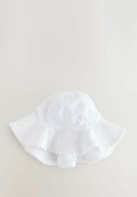 Valkoinen ämpärilakki, joka on valmistettu teksturoituneesta kankaasta ja jossa on kukkakuviointia. Lakin reunassa on röyhelö ja pomponkoriste.