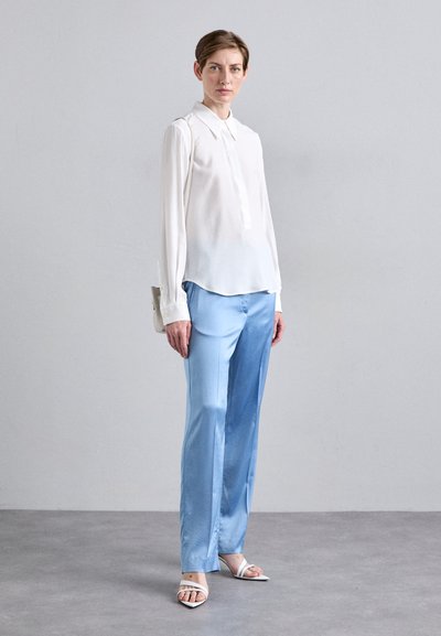 White long-sleeve blouse with a button-down collar, paired with light blue satin trousers. Model wears white strappy sandals. Simple bag.