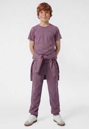 Niño pelirrojo sonriente, vestido con una camiseta de manga corta morada, pantalones a juego, una sudadera atada a la cintura y zapatillas blancas.