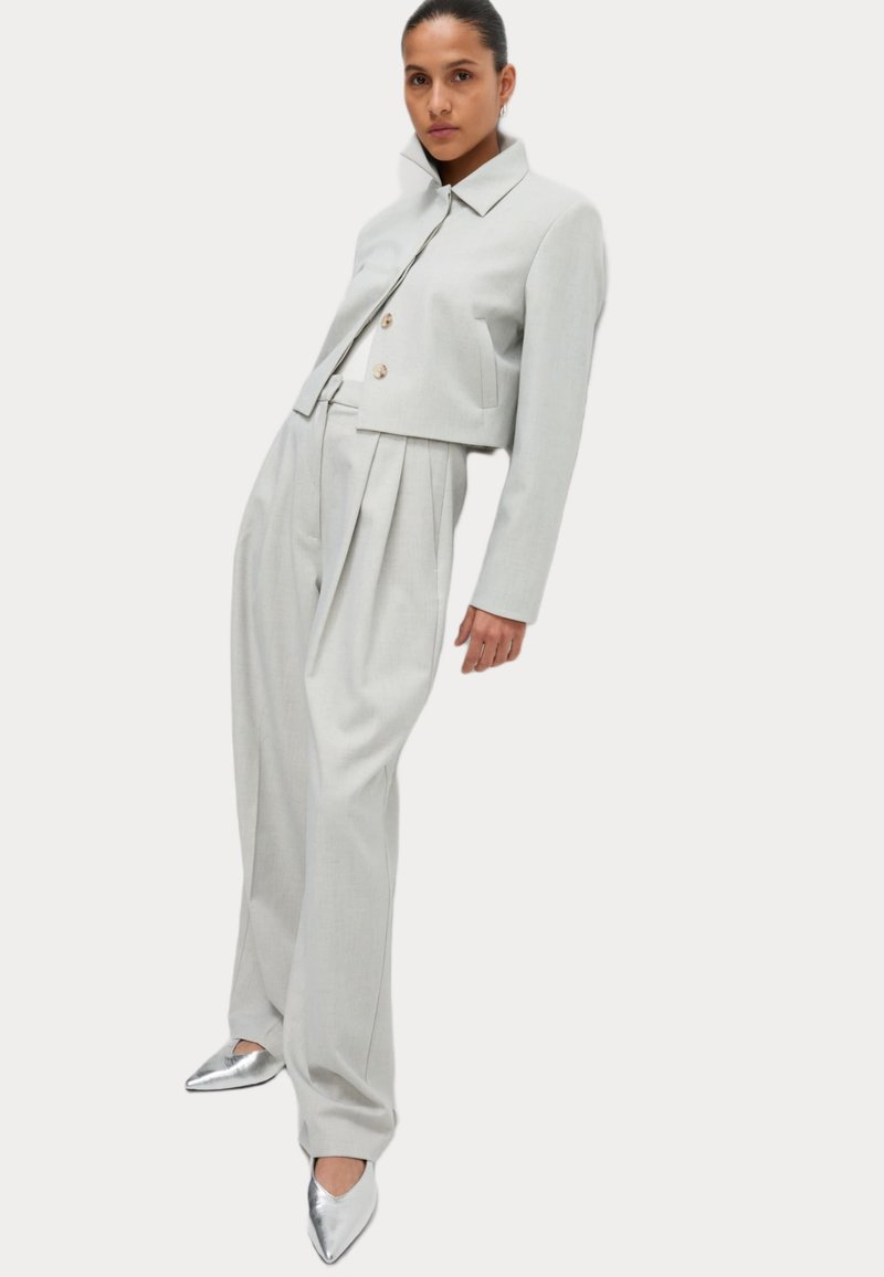 Ljust grå skräddarsydd kostym med en kort jacka och högmidjade veckade byxor. Silverfärgade klackskor med spetsig tå kompletterar outfiten.
