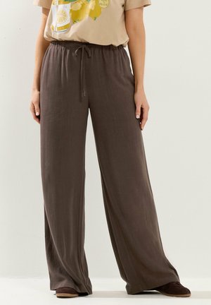 Osoba w luźnych, brązowych spodniach o wysokim stanie i szerokich nogawkach z troczkiem oraz brązowych butach, w połączeniu z beżowym t-shirtem z nadrukiem cytryny.