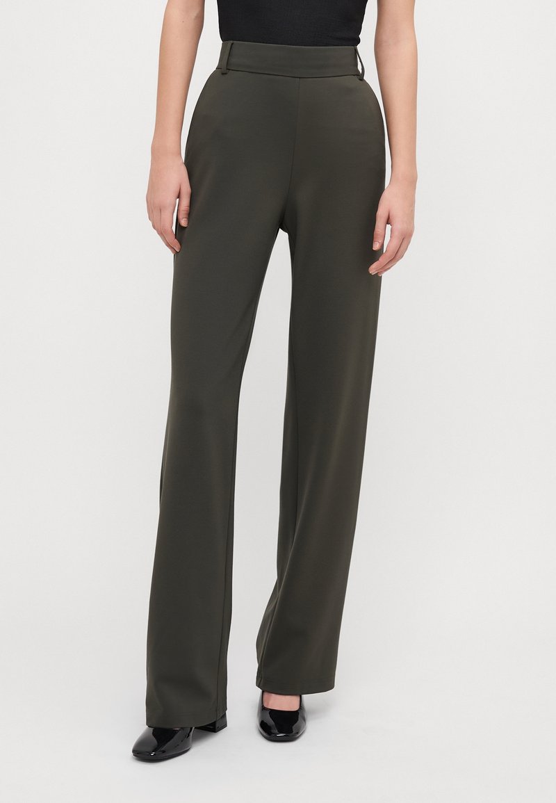 Femme portant un pantalon large taille haute de couleur olive foncée et des chaussures à talons noires brillantes, debout devant un fond clair uni.