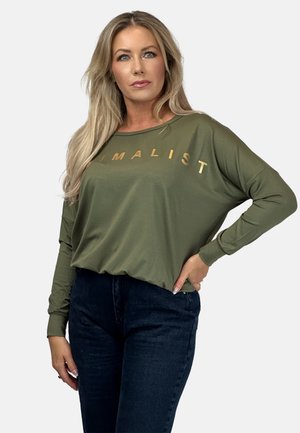 Blondynka z długimi włosami, ubrana w oliwkową koszulę z długim rękawem z napisem "MINIMALIST" w złotych literach oraz ciemnoniebieskie dżinsy, z jedną ręką na biodrze.