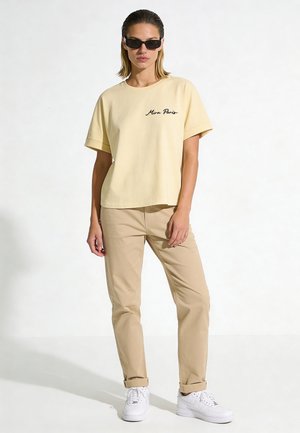 Vrouw met beige T-shirt met tekst, kaki broek, witte sneakers en zwarte zonnebril staand tegen een witte achtergrond.