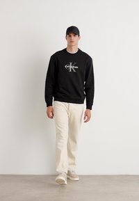 Sweatshirt noir avec le logo "Calvin Klein", pantalon crème et baskets grises. Design simple, texture lisse et coupe décontractée.
