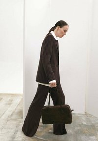 Completo marrone a righe sottili con pantaloni a gamba larga e blazer a bottone singolo. Camicia bianca con colletto sotto. Tiene una borsa rettangolare marrone scuro.