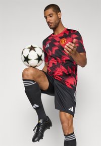 Rot und schwarz gemustertes Fußballtrikot mit dem Manchester United Logo, schwarze Shorts, schwarze Socken mit weißen Streifen und schwarze Fußballschuhe.