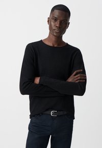 Czarny sweter z długim rękawem i okrągłym dekoltem, wykonany z miękkiego materiału, noszony z ciemnymi dżinsami i czarnym paskiem, z rękami skrzyżowanymi na przedzie.