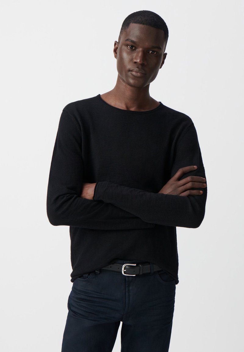 Czarny sweter z długim rękawem i okrągłym dekoltem, wykonany z miękkiego materiału, noszony z ciemnymi dżinsami i czarnym paskiem, z rękami skrzyżowanymi na przedzie.