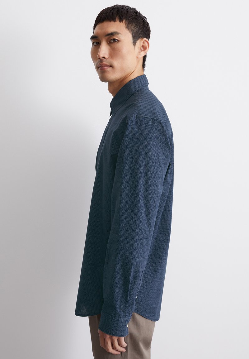 Chemise à manches longues, bleu marine, en tissu texturé, avec col boutonné et fentes latérales. Présente une coupe classique et des poignets à boutons.