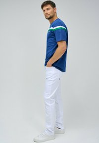 Camiseta de algodón azul con detalles en rayas blancas y verdes, combinada con pantalones de algodón blancos y zapatillas blancas, de pie frente a un fondo simple.