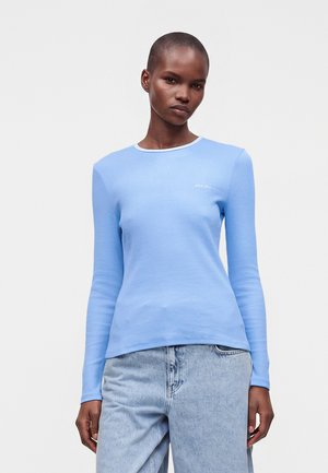 Femme portant une chemise ajustée bleu clair à manches longues et un jean bleu clair, debout devant un fond clair uni.