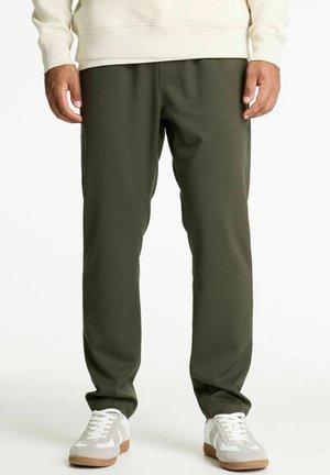 Homme portant un pantalon casual vert olive, des baskets blanches avec des accents beiges, et un sweatshirt crème, se tenant sur un fond blanc.