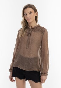 Brun mønstrede bluse med gennemsigtigt stof, flæset halsudskæring og lange ærmer med elastiske manchetter, parret med sorte shorts.