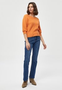 Maglione arancione lavorato a maglia con maniche a tre quarti, abbinato a jeans dritti blu e stivali alla caviglia beige. Design semplice e texturizzato.