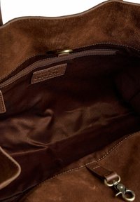 Interno della tote bag in suede marrone con una tasca con zip, fodera in tessuto morbido e un piccolo tag in pelle con il nome del brand.
