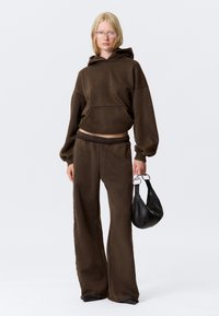 Bruine hoodie met een cropped ontwerp, gecombineerd met bijpassende wijde sweatpants. Model houdt een kleine zwarte handtas vast.