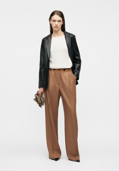 Femme debout portant un blazer en cuir noir, un haut blanc, un pantalon large marron, des chaussures noires, et tenant une pochette à imprimé léopard dans sa main droite.
