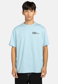 Camiseta de algodón azul claro con mangas cortas. Presenta un logo impreso en negro en el lado izquierdo del pecho. Corte relajado y diseño simple.