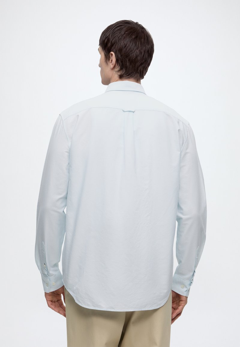 Uomo con camicia azzurro chiaro a maniche lunghe e pantaloni beige, in piedi di spalle alla macchina fotografica contro uno sfondo chiaro e semplice.