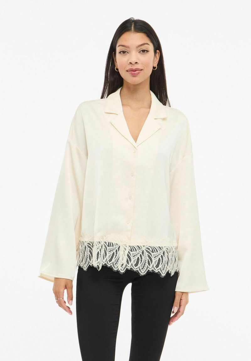 Kremfarget silkeskjorte med knappestolpe foran, hakket krage og vide ermer. Har blondekant ved kanten og glatt tekstur.