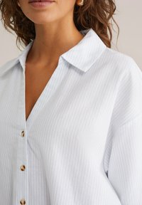 Light blue and white striped shirt with a collar, button front, and loose fit. Cotton material with a soft texture and subtle sheen.
