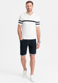 Camisa polo blanca con rayas marinas, combinada con pantalones cortos de mezclilla oscuros, con dobladillo a la altura de las rodillas, y zapatillas blancas.