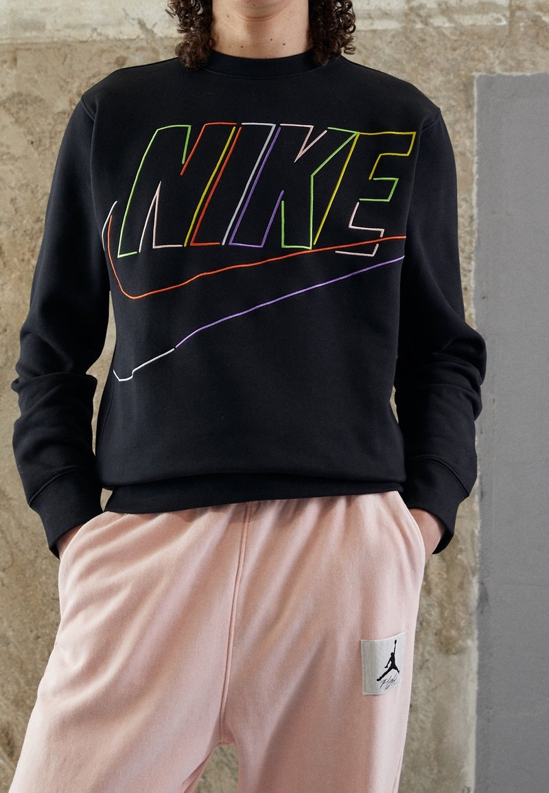 Person klädd i svart sweatshirt med flerfärgad NIKE-logga och rosa byxor med Jordan-logga, stående mot en strukturerad vägg.