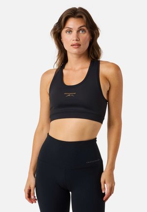 Donna che indossa un reggiseno sportivo nero e leggings a vita alta, in piedi di fronte a uno sfondo bianco, con un'espressione rilassata.