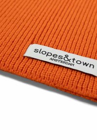 Tessuto arancione a costine con un'etichetta bianca che recita "slopes&town AMSTERDAM," con testo nero. Il tessuto ha un aspetto testurizzato.