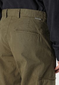 Oliivroheline cargo-püksid, millel on küljetaskud, lüliga vöökoht ja kerge sära. Kangas paistab sileda ja peene tekstuuriga.