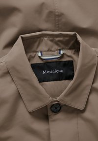 Beež mantli krae, millel on üks must nupp, triibuline kangasrõngas ja must silt, millel on kaelas sees kiri "Matinique".