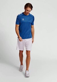 Modré sportovní tričko s bílými detaily, kulatým výstřihem, krátkými rukávy a logem; spárováno s bílými šortkami a světlými teniskami.