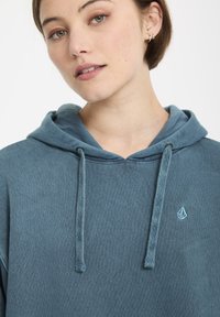 Hoodie in diepblauw met een gestructureerde afwerking, voorzien van een centrale trekkoord en een klein geborduurd logo op de borst.