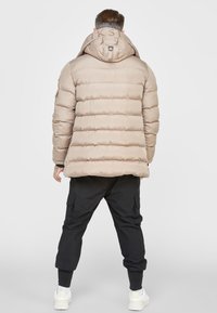 Giacca trapuntata beige con cuciture orizzontali, cappuccio e dettagli ricamati sul collo, abbinata a pantaloni cargo neri e sneakers bianche.