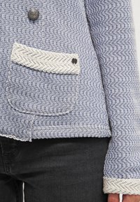 Blå och vit texturerad kavaj med en ficka med vävd kant, en dekorativ knapp och fransade kanter vid ärmarnas fåll.