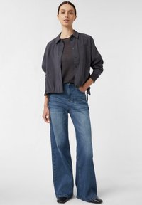 Graues Hemd mit Knöpfen, graues T-Shirt darunter, blaue weite Jeans, schwarze Schuhe. Material wirkt leicht und hat eine glatte Textur.