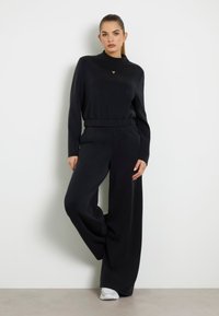 Čierny dvojdielny outfit pozostávajúci z blúzky s dlhým rukávom a stojačikom, s malým trojuholníkovým logom, v kombinácii s nohavicami so širokými nohavicami. Hladká textúra.