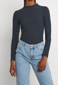 Mörkgrå långärmad ribbad tröja med vågig halsringning, ihop med högmidjade ljusblå denimjeans med klassisk passform och knäppning.