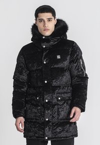 Čierna nadýchaná parka vyrobená z textúrovaného materiálu, s kapucňou podšitou kožušinou, patentkami, bočnými vreckami a vyšitým logom.