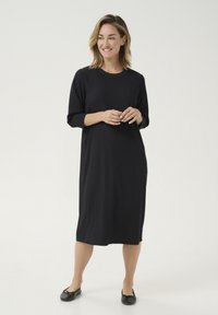 Robe noire à manches trois-quarts avec une coupe ample et une surface texturée, arrivant au milieu du mollet. Portée avec des chaussures plates noires.