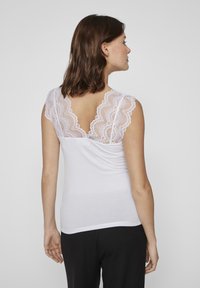 Wit tanktop met kantdetails op de schouders en rug, met een getailleerd ontwerp en ribbelstructuur. Zwarte broek zichtbaar onderaan.