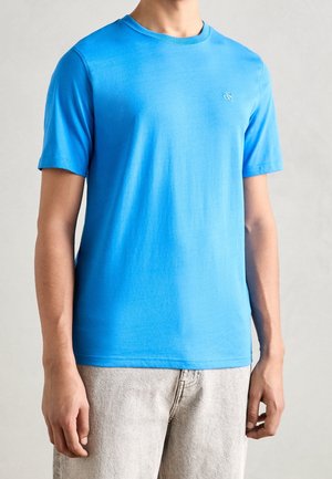 Mężczyzna ubrany w gładki, jasnoniebieski t-shirt z krótkim rękawem oraz jasnobeżowe spodnie na neutralnym tle.