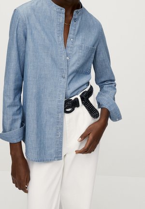 Personne à la peau foncée portant une chemise bleu clair sans col à boutons avec les manches retroussées, un pantalon blanc et une ceinture tressée noire.