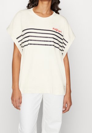 Kobieta ubrana w luźny kremowy T-shirt z czarnymi poziomymi paskami i czerwonym napisem „Hilfiger”, zestawiony z białymi spodniami.