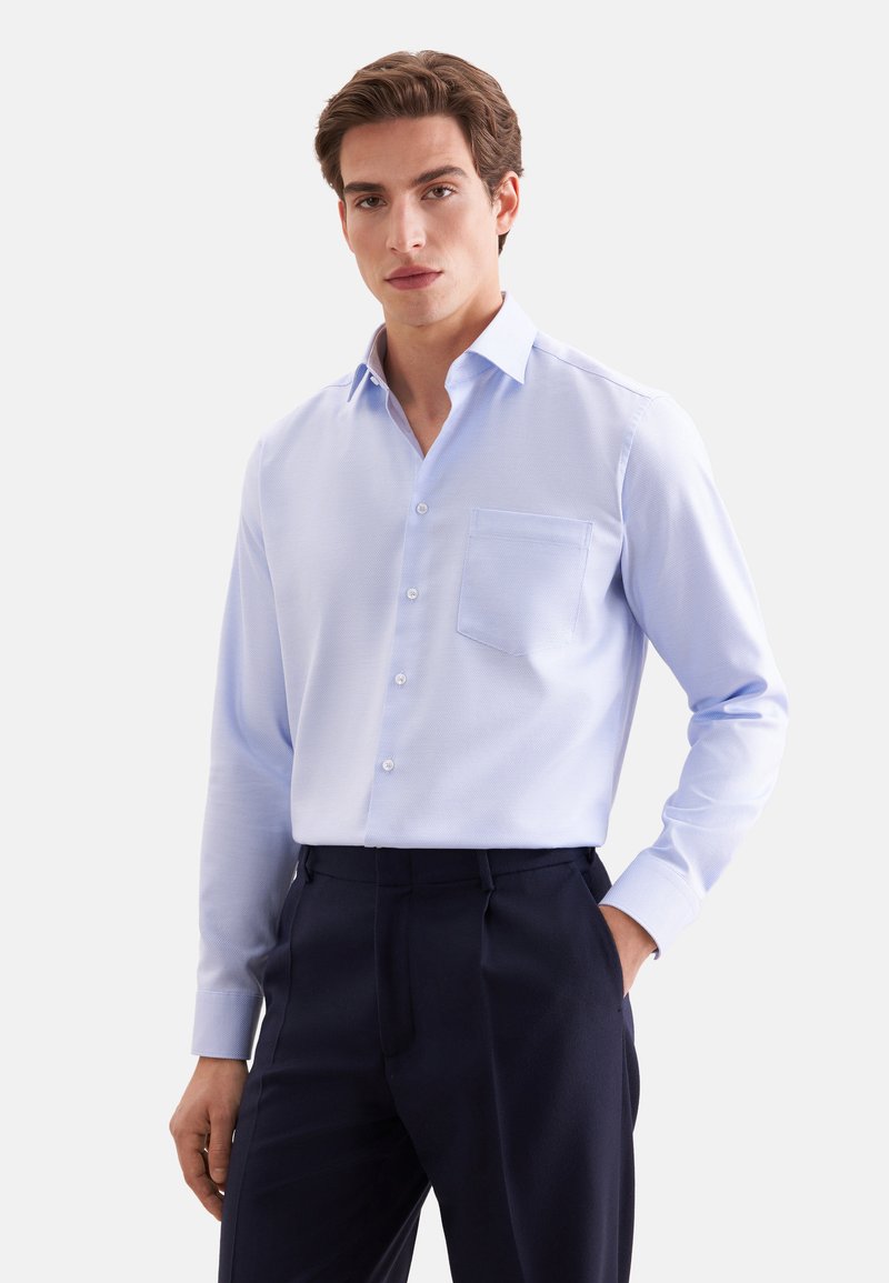 Homme portant une chemise bleu clair boutonnée avec une poche poitrine et un pantalon foncé, debout avec une main dans la poche.